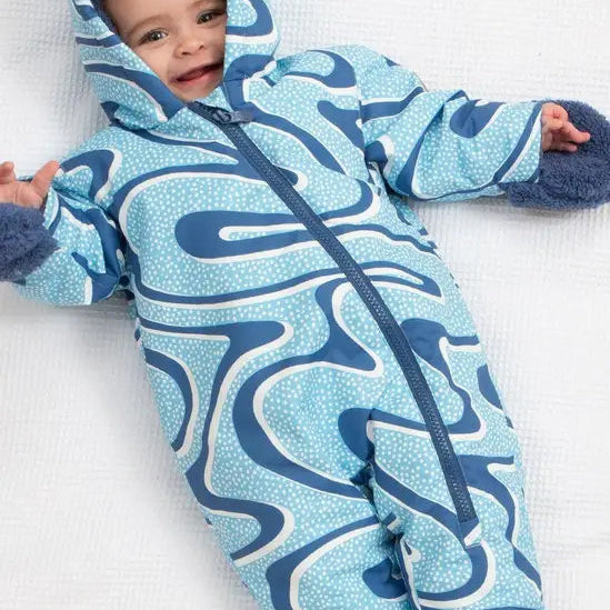 A baby lying on a white textured blanket, wearing a hooded one-piece snowsuit with a blue, navy, and white wavy pattern.