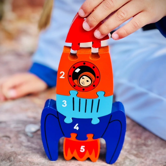 A colorful wooden rocket-shaped toy being held by a child's hand outdoors. The toy has numbered sections from 1 to 5 and features a cartoon astronaut face in the center.