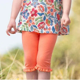 Child wearing a floral top and orange pants standing in a grassy field.