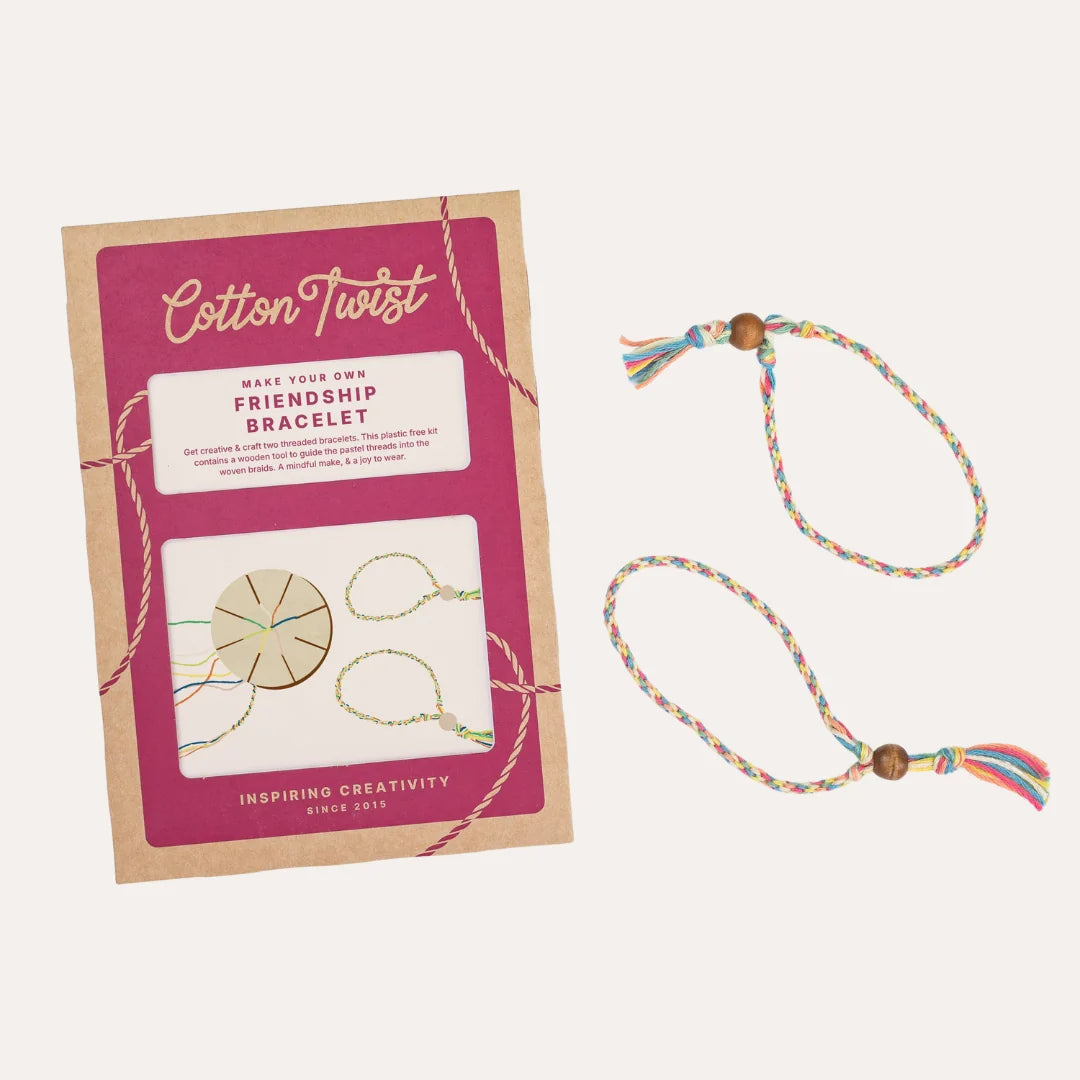 A friendship bracelet kit by Cotton Twist, featuring a kraft paper package with a magenta label and two completed multicolor braided cord bracelets with wooden beads and tassels, displayed on a light cream background.