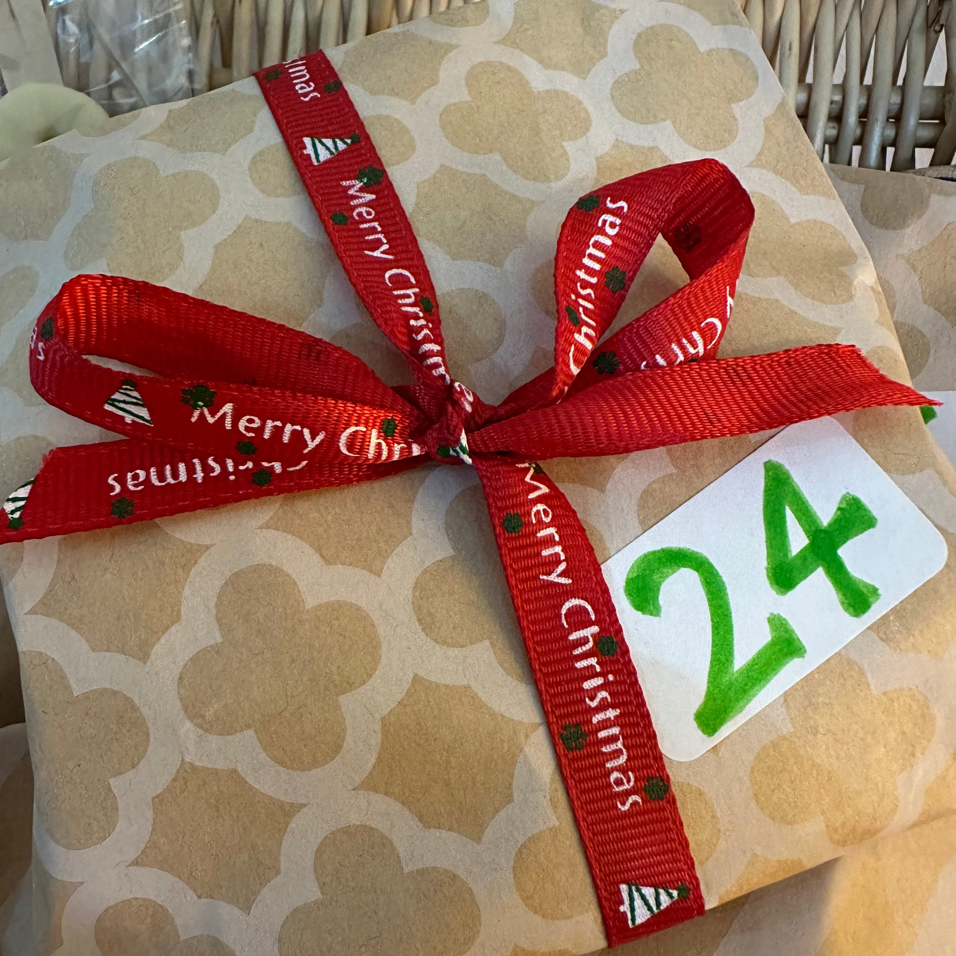 A wrapped gift box with beige patterned paper, tied with a red ribbon printed with 'Merry Christmas' and a white tag showing the number 24.
