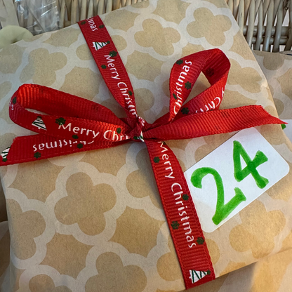 A wrapped gift box with beige patterned paper, tied with a red ribbon printed with 'Merry Christmas' and a white tag showing the number 24.