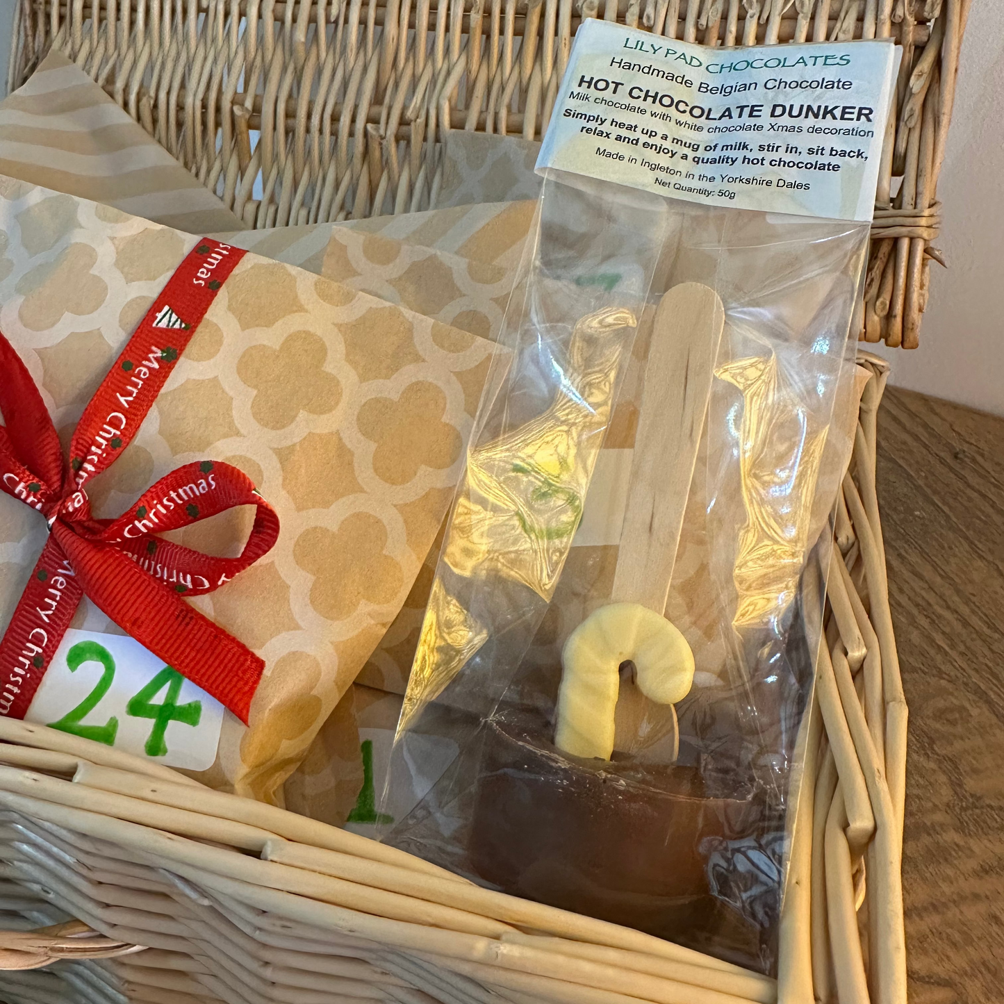 A wicker gift basket filled with treats, including a packaged hot chocolate dunker in a clear bag with a wooden stick and a yellow candy accent, next to a wrapped present tied with a red ribbon and a white tag reading 24.