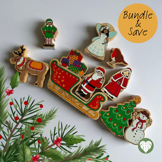 A set of colorful wooden Christmas figurines arranged on a white surface with decorative pine branches and berries. The set includes a reindeer, Santa Claus in a sleigh with gifts, an angel, a rooster, an elf, a Christmas tree, a snowman, and a Santa-like figure.
