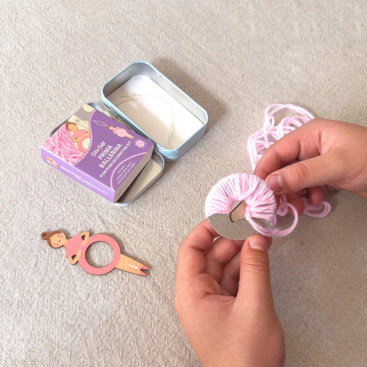 A person's hands are shown wrapping light pink yarn around a silver metal ring, likely making a hair tie or hair accessory. A purple tin of 'Sofia's Saltirama' brand yarn is visible in the background, along with a small wooden ballerina hair tie tool on a textured beige fabric surface.