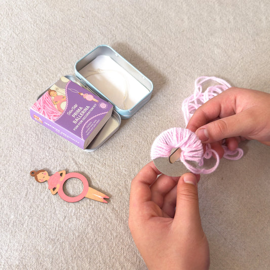 A person's hands are shown wrapping light pink yarn around a silver metal ring, likely making a hair tie or hair accessory. A purple tin of 'Sofia's Saltirama' brand yarn is visible in the background, along with a small wooden ballerina hair tie tool on a textured beige fabric surface.