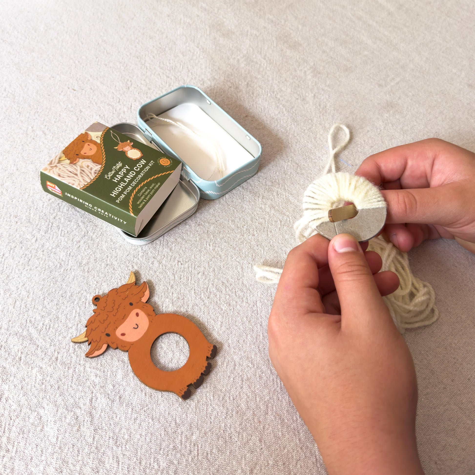 A person's hands are shown wrapping cream-colored yarn around a wooden ring-shaped teether. The product set includes the wooden teether with a cow face, a small green and gold tin box, and an open metal tin containing the yarn.