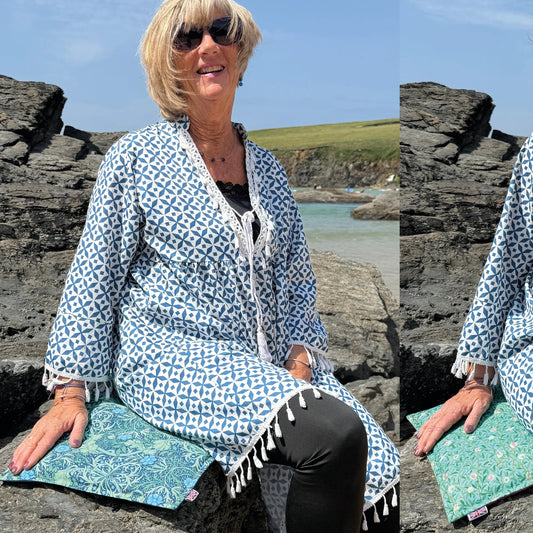 A woman sitting outdoors on rocks by the sea, wearing a blue and white geometric patterned cardigan or cover-up over a black top and black leggings. She is also wearing sunglasses and holding a teal patterned pillow.