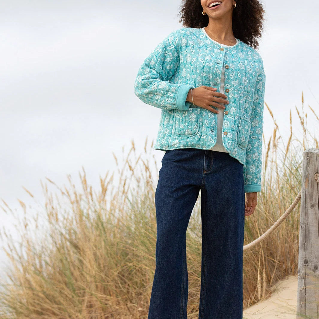 Woman wearing a light blue patterned jacket and dark blue jeans standing on a sandy path with grasses in the background.