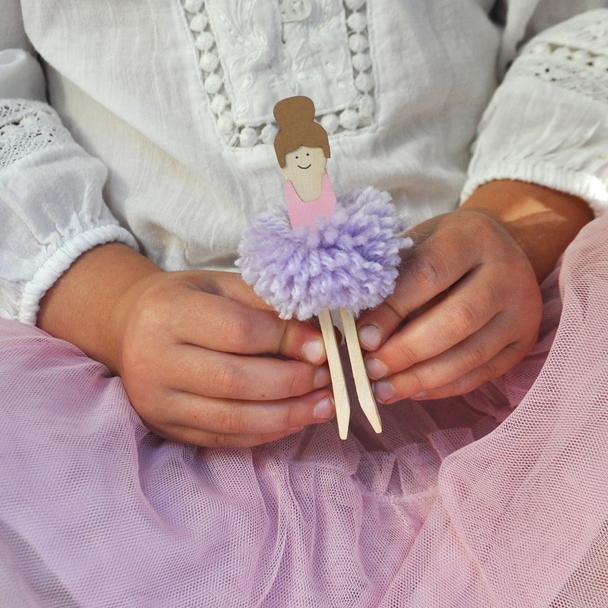 A close-up of a child's hands holding a handmade wooden ballerina clipper. The clipper features a wooden figure with a brown hair bun, a pink dress, and a fluffy light purple pom-pom tutu, attached to two wooden legs.
