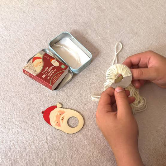 A person's hands are shown holding a white yarn-covered Christmas ornament with a Santa face cutout, next to an open tin of Santa ornaments and a Santa ornament on a textured beige fabric background.