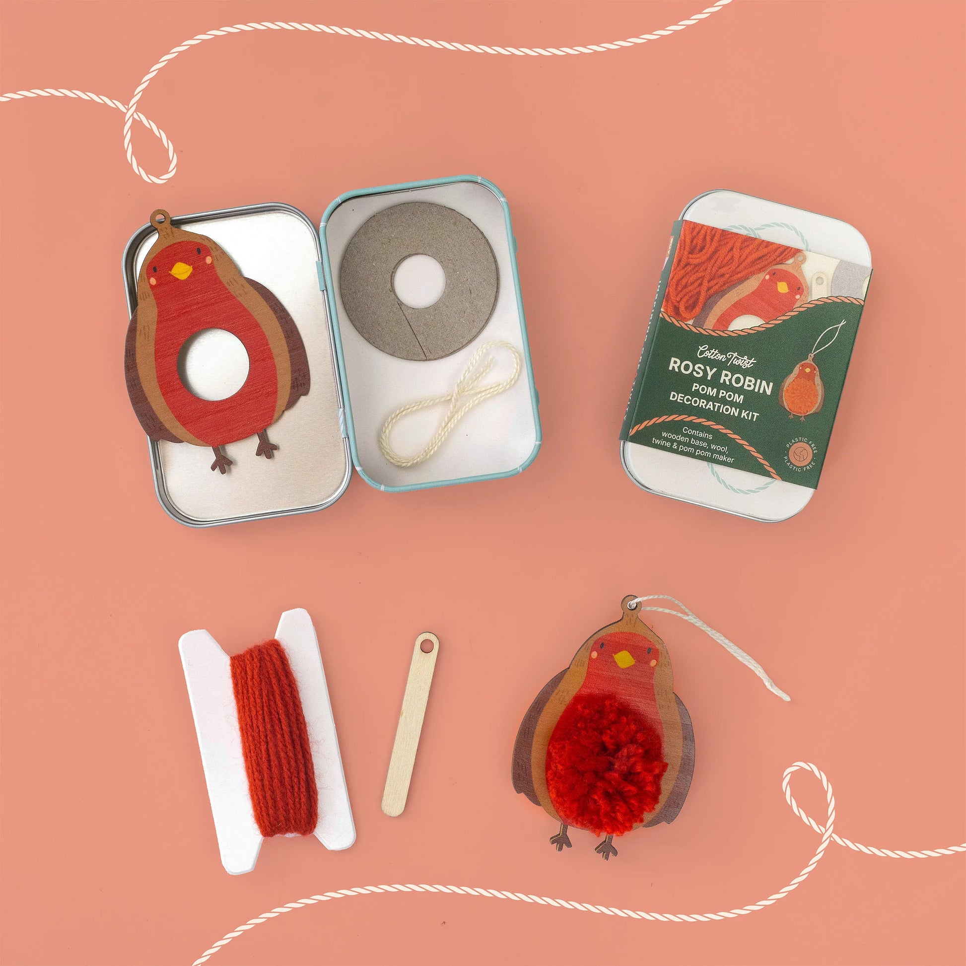 A DIY Rosy Robin decoration kit laid out on a peach background, including a tin with a felt robin decoration, a spool of red yarn, a wooden stick, and another packaged robin decoration in the background.