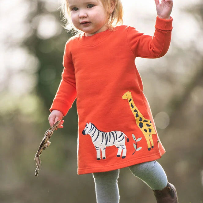 A toddler girl wearing a bright orange long-sleeve dress with a zebra and giraffe graphic, standing outdoors on a grassy lawn.