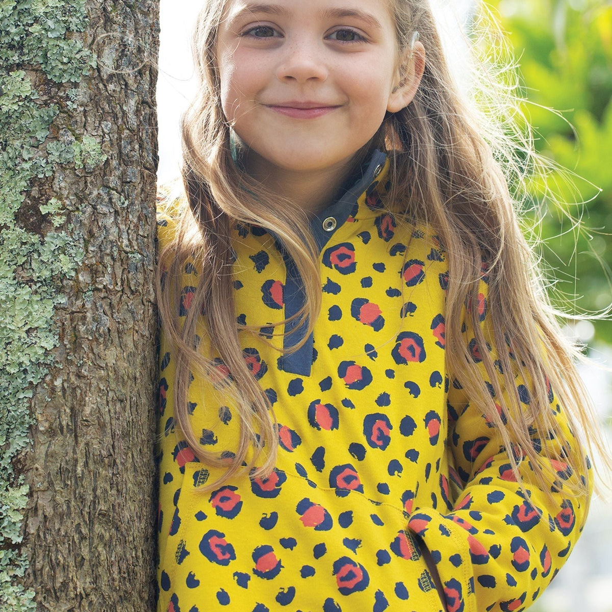 A young girl with long light brown hair smiles while leaning against a tree trunk outdoors. She is wearing a bright yellow fleece pullover with a navy blue collar and a red and navy leopard print pattern.