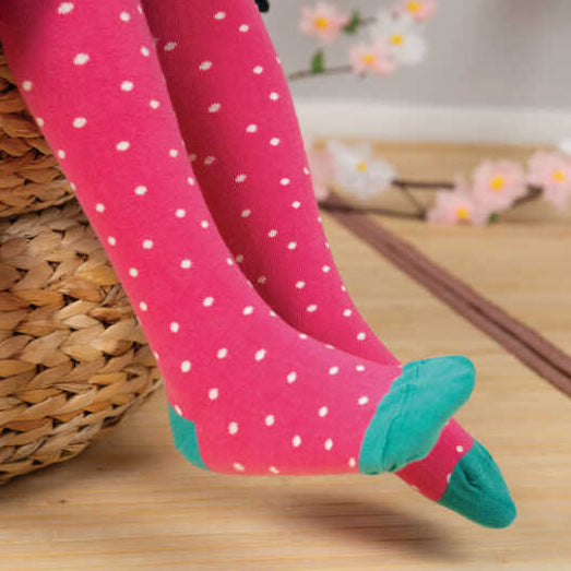Pink tights with white polka dots and teal toe/heel accents, worn by a child seated on a woven basket with a light wood floor and soft pink flowers in the background.