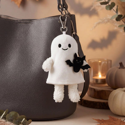 A white plush ghost keychain charm with a black embroidered face and a small black bat accessory, hanging from a dark gray textured handbag in a warm autumn-themed setting with pumpkins, leaves, and a candle.