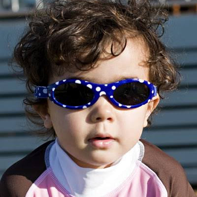 A close-up of a toddler with curly brown hair wearing blue and white polka dot swim goggles and a pink and brown swim shirt against a blue siding background.
