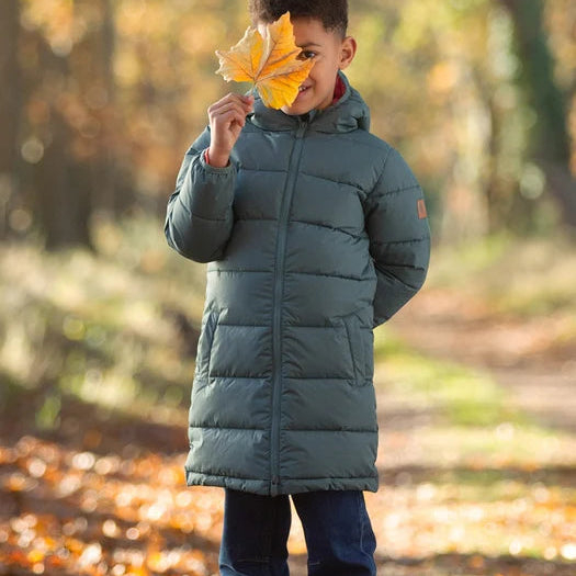 A child wearing a long, dark teal-green puffer parka and blue jeans stands on a leaf-covered path in an autumn forest, holding a yellow leaf in front of their face.