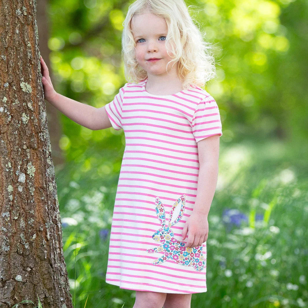 A young girl with blonde hair stands outdoors next to a tree, wearing a pink and white striped short-sleeve dress with a colorful bunny applique, pink socks, and white sneakers.