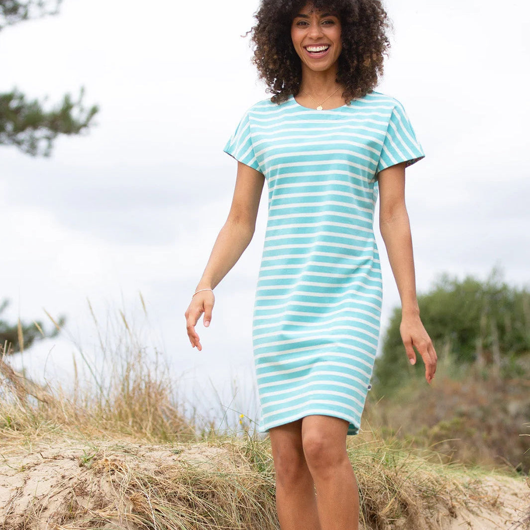 A woman wearing a short-sleeve, knee-length striped dress in mint green and white stands on a sandy dune outdoors under an overcast sky.