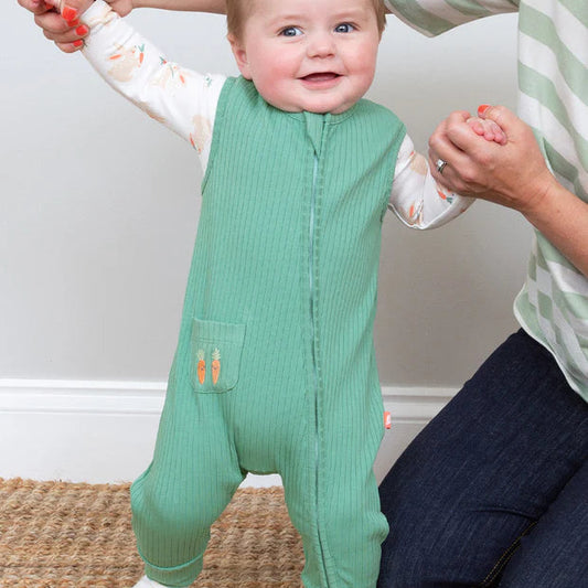 Baby in a green onesie standing with a person holding their hands on a carpeted floor.