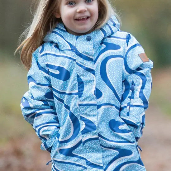 A young girl with blonde hair smiling outdoors, wearing a light blue hooded jacket with a bold dark blue abstract wavy pattern and dark green pants.