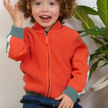 A smiling young child with curly brown hair sits on a wooden bench wearing a bright orange ribbed zip-front sweater with sage green and white striped cuffs, paired with blue jeans. A green houseplant is visible in the background against a light wall.