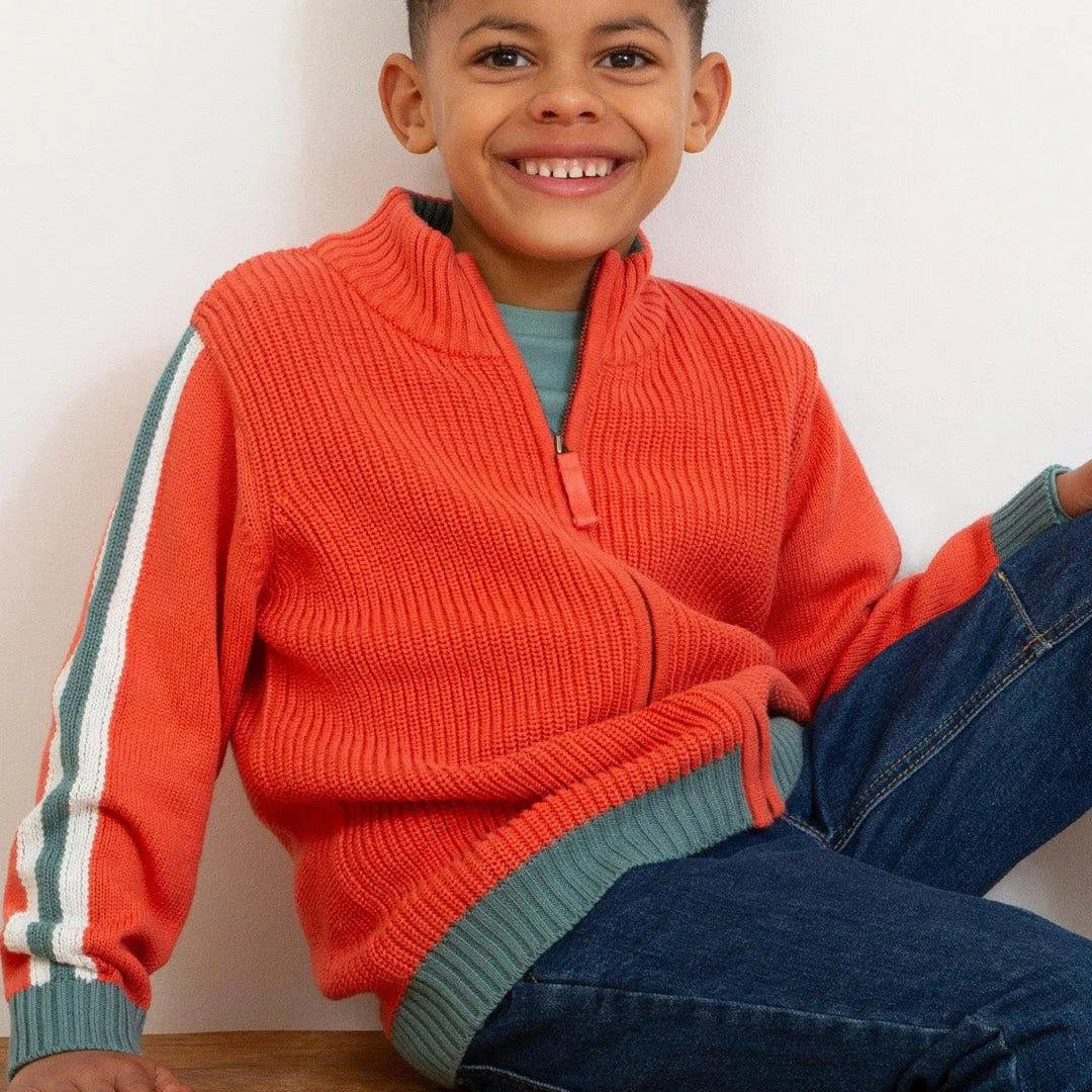 A smiling young boy wearing a bright orange ribbed quarter-zip sweater vest over a teal shirt and blue jeans, sitting on a wooden bench against a plain white wall.
