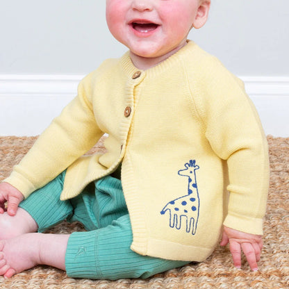 Baby wearing a yellow cardigan with a giraffe design, sitting on a textured surface.