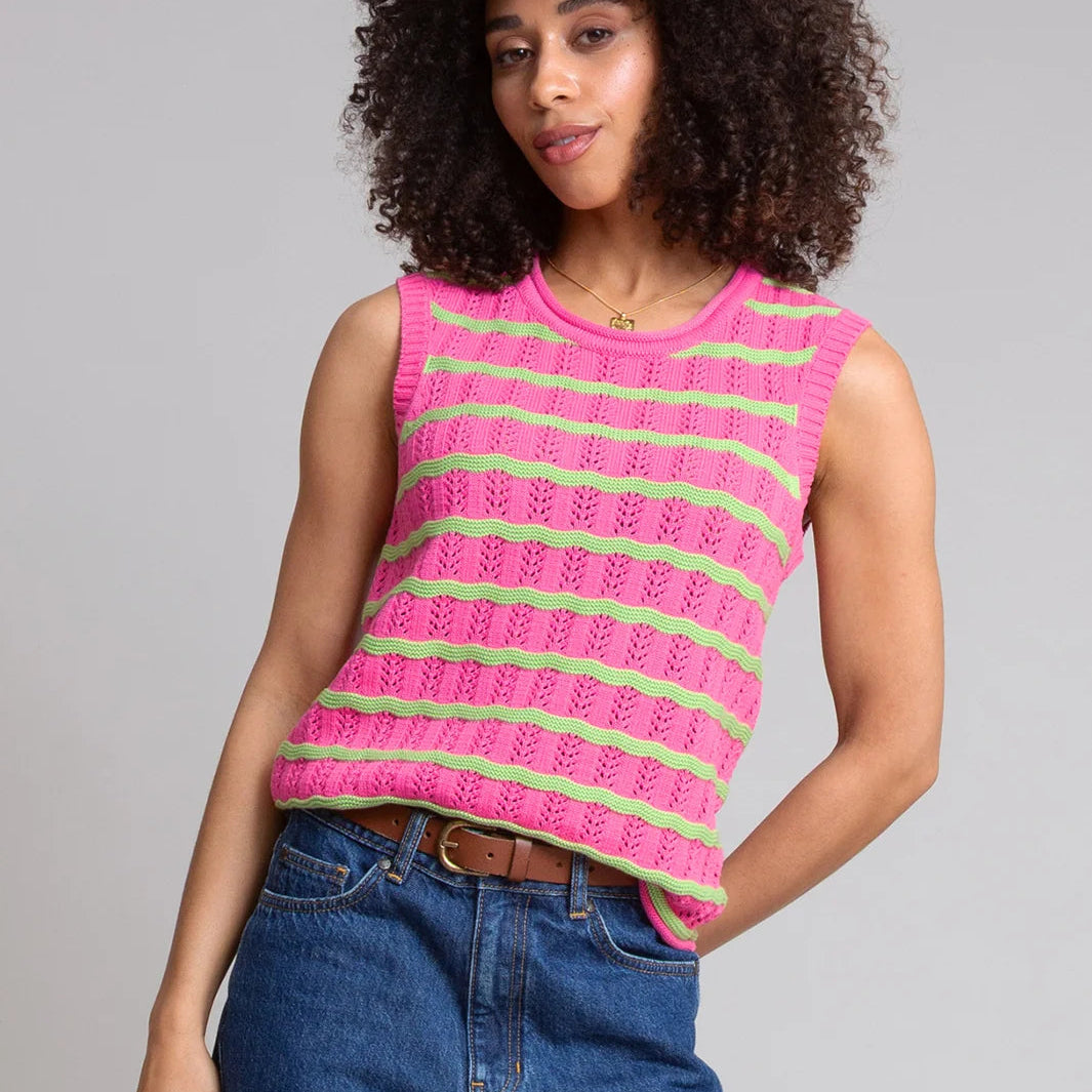 A woman with curly dark hair wearing a bright pink sleeveless knit top with lime green horizontal stripes, paired with blue jeans, against a solid gray studio background.