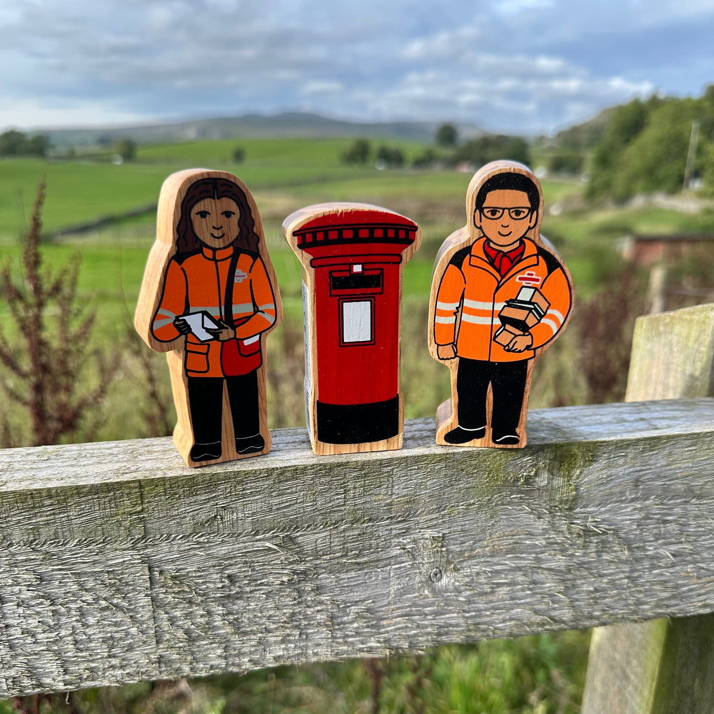 Three hand-painted wooden toy figures representing a postman and two workers in orange high-visibility jackets, standing on a weathered wooden fence in a rural outdoor setting.