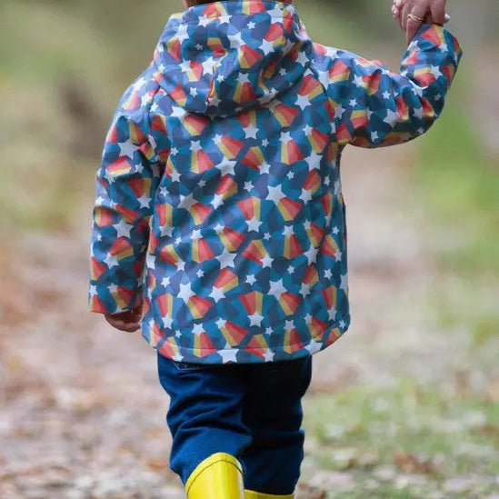 A child seen from behind wearing a hooded rain jacket with a colorful candy corn and star pattern, blue pants, and yellow rain boots while walking outdoors on a path and holding an adult's hand.