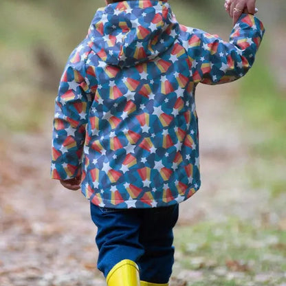 A child seen from behind wearing a hooded rain jacket with a colorful candy corn and star pattern, blue pants, and yellow rain boots while walking outdoors on a path and holding an adult's hand.