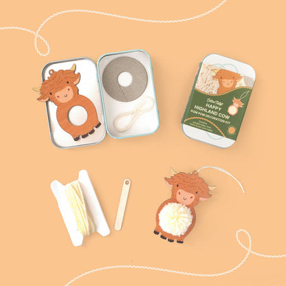 A flat-lay view of a 'Happy Highland Cow' pom-pom craft kit on a peach background. The kit includes a small metal tin with a cow-shaped ornament, a roll of gray pom-pom string, a white cord, and a green cardboard backing card.