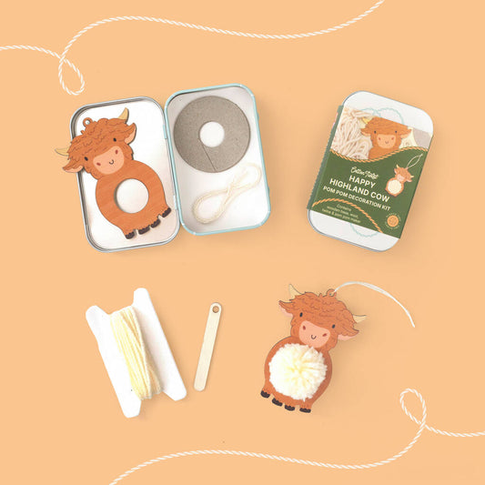 A flat-lay view of a 'Happy Highland Cow' pom-pom craft kit on a peach background. The kit includes a small metal tin with a cow-shaped ornament, a roll of gray pom-pom string, a white cord, and a green cardboard backing card.