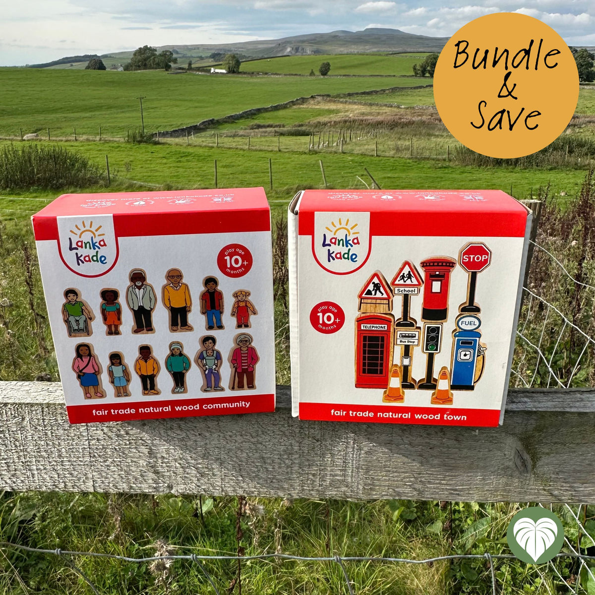 Two Lanki Kade toy boxes displayed outdoors on a wooden fence with a green rural landscape in the background. One box shows wooden community figures, and the other shows red traditional British road and telephone accessories.