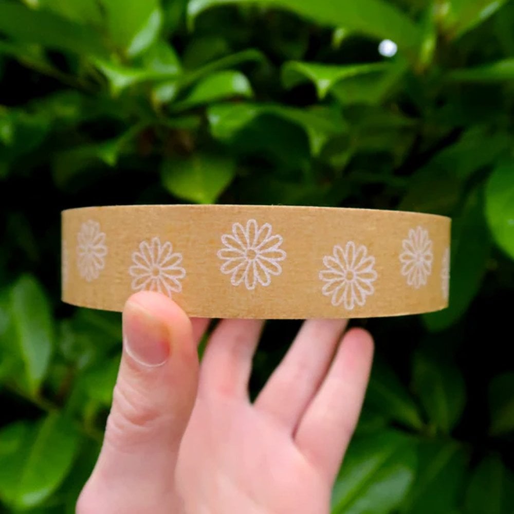 A hand holding a brown kraft paper band decorated with a repeating white daisy pattern, set against a background of green leaves.